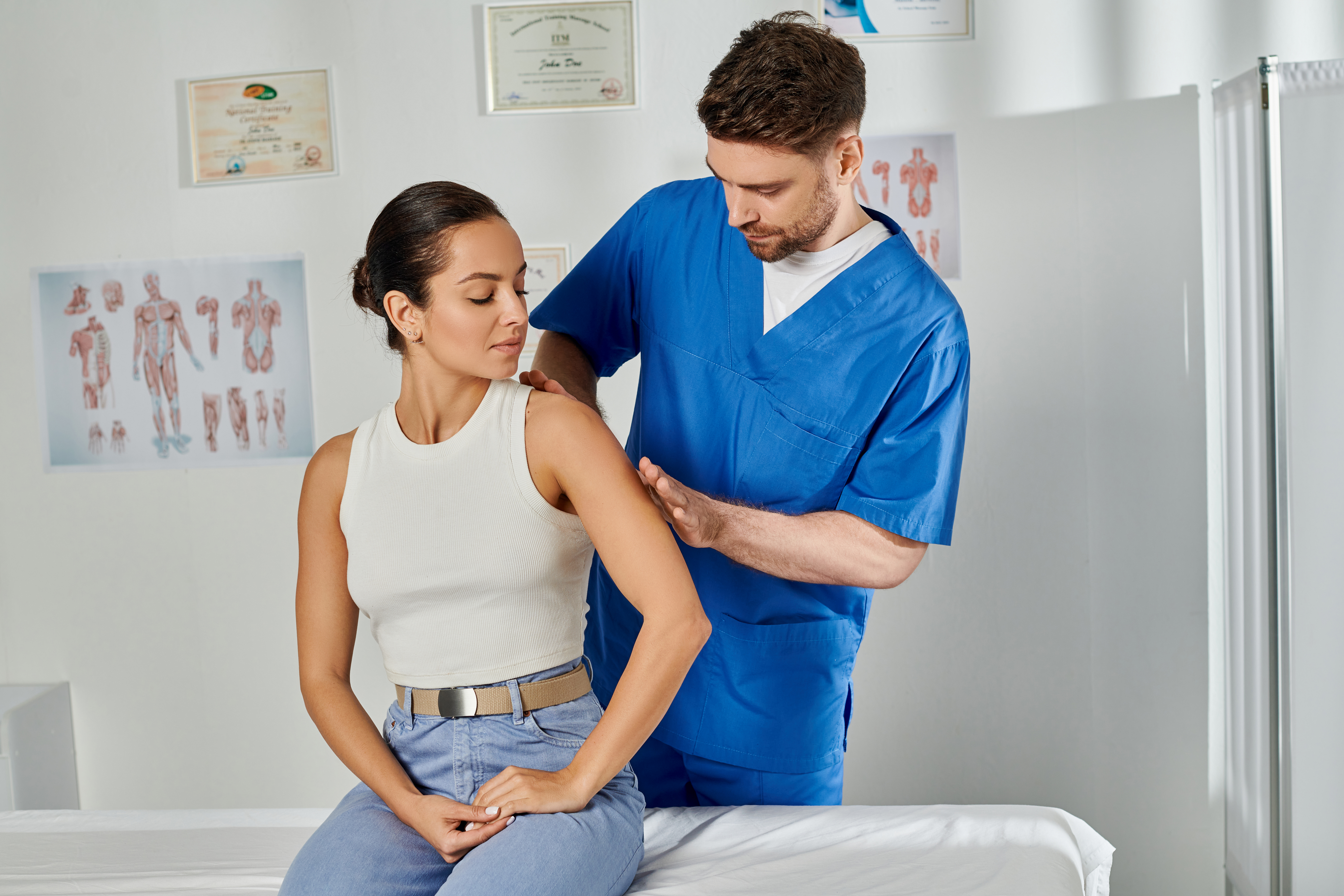 handsome doctor in blue medical costume massaging shoulder of his young attractive patient