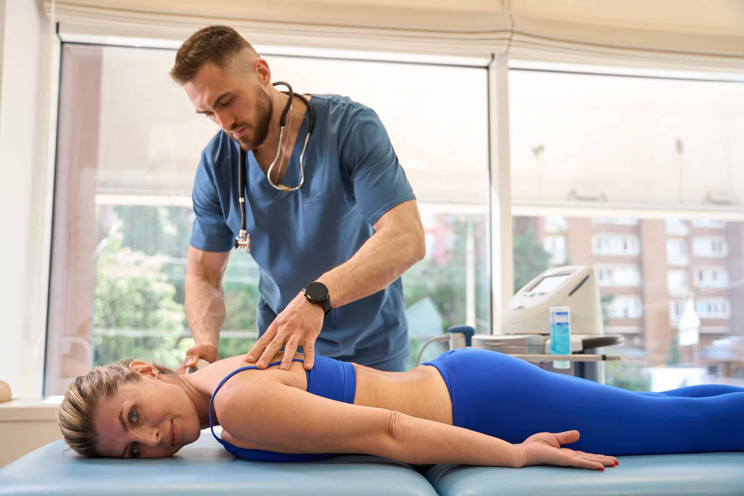 Woman patient lying on massage couch while therapist doing extracorporeal shock wave therapy affecting on trigger points on her body, treatment of various pains of musculoskeletal system