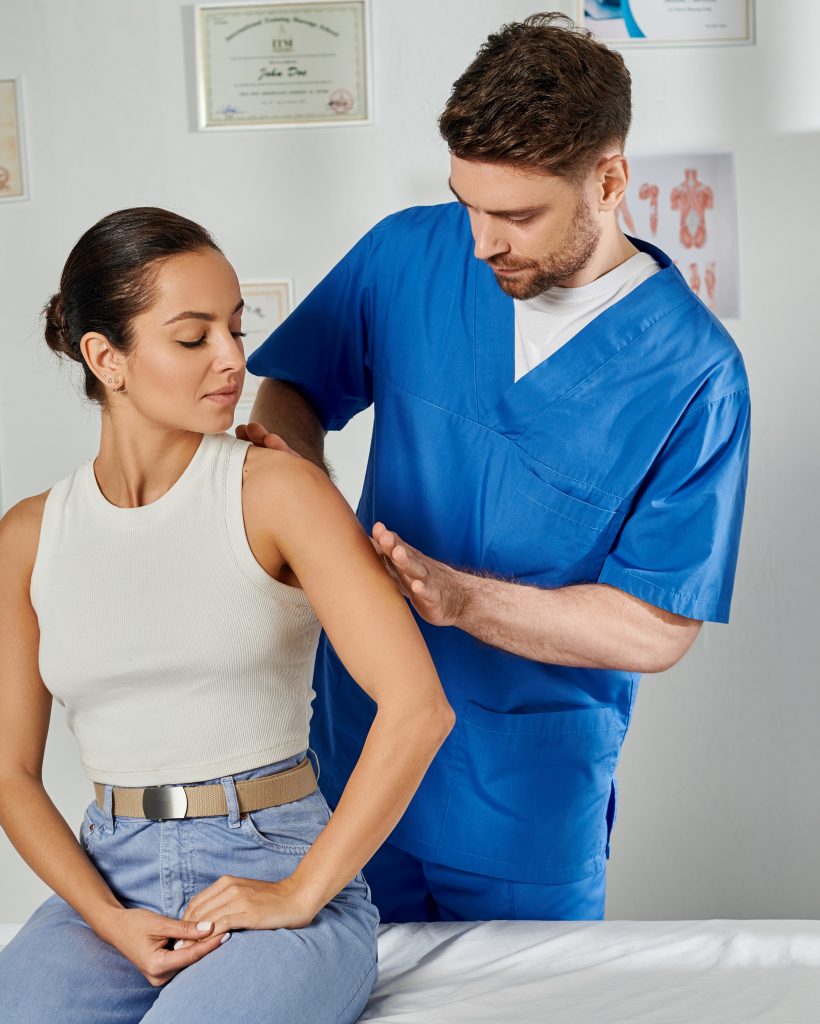 handsome doctor in blue medical costume massaging shoulder of his young attractive patient